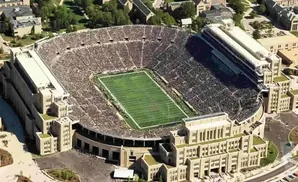 Aerial view of a packed football stadium with a green field, surrounded by stone buildings and trees.