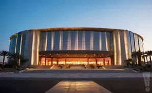 Modern, glass-fronted circular building with illuminated columns and palm trees outside.