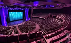 An empty theater with a stage and tiered seating illuminated by purple and blue lights.