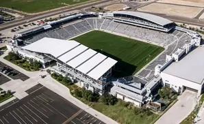 Aerial view of a stadium with a green field, tiered seating, and white-roofed buildings.