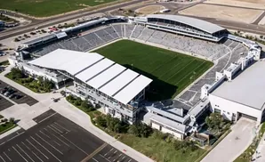 Aerial view of a modern stadium with a green field, tiered seating, and white-roofed buildings.