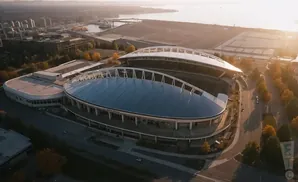 Aerial view of a stadium with a blue roof, surrounded by trees and buildings, at sunset.