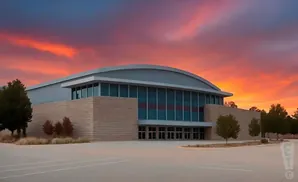 A modern building with a curved roof and large windows stands under a vibrant orange and red sunset sky.
