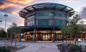 Modern circular building with a glass roof and large windows, with trees and benches in front during sunset.