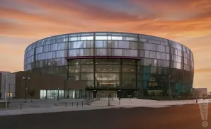 A modern circular glass arena at sunset with an orange sky.