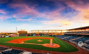 A baseball stadium during a colorful sunset, with a game in progress on the field.