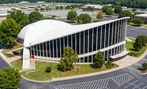Aerial view of a modern building with a curved white roof and large glass windows, surrounded by trees and parking lots.