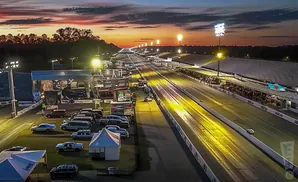 A drag strip at sunset with lights illuminating the track and stands filled with spectators.