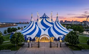 A large blue and white striped circus tent at dusk, with a Ferris wheel and city skyline in the background.