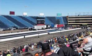 Spectators at a drag race in a stadium, with blue bleachers and a race track visible under a clear blue sky.