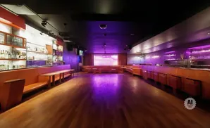 A dimly lit nightclub with purple lighting, featuring a long bar, booths, and a stage with a screen.