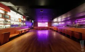 An empty, dimly lit nightclub with orange booths, a bar, and a large projection screen.