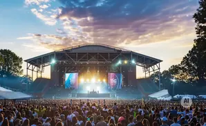 A large crowd watches a concert on an outdoor stage at sunset, with bright lights and large screens.