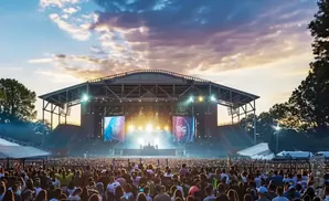 Outdoor concert at dusk with a large crowd facing a stage and screens under a cloudy sky.