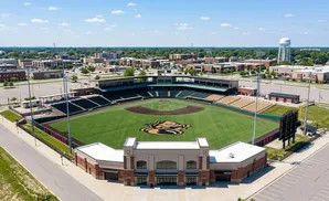 Aerial view of a baseball stadium with green field, stands, and surrounding buildings under a blue sky with clouds.
