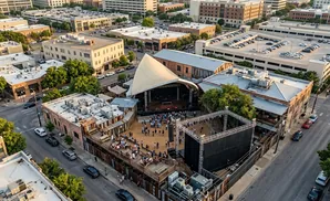 Aerial view of an outdoor concert venue with a large stage and seating areas, surrounded by urban buildings and parking garages.