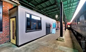 A modern gray building with a yellow door and large window sits beside a historic train on a platform.