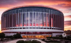 A modern stadium reflects a colorful sunset under a dramatic sky.