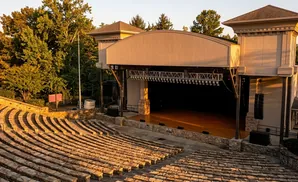 Outdoor amphitheater with stone seating and a stage under a covered pavilion, bathed in warm, golden sunlight.