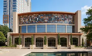 Building with a mosaic mural above large windows, flanked by trees and a pool.