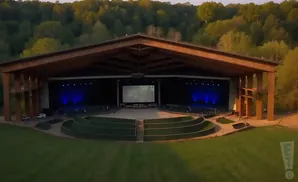 Outdoor amphitheater with a covered stage, screen, and tiered seating surrounded by grass.