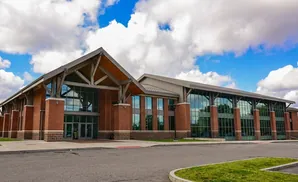 Modern brick building with large glass windows and a wooden-beamed pitched roof against a cloudy blue sky.