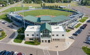 Aerial view of a baseball stadium with a main building, bleachers, and a parking lot filled with cars.