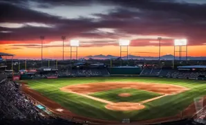 A baseball stadium at sunset with lights on and a colorful sky over distant mountains.