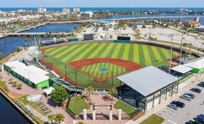 Aerial view of a baseball field with green and red infield, surrounded by buildings, a parking lot, and a body of water.