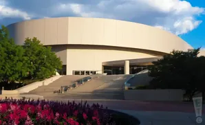 A modern, curved building with a wide staircase leading to its entrance, surrounded by trees and flowers.