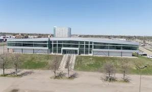 Modern glass-fronted office building with stairs leading to the entrance, surrounded by trees and parking lots.