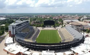 Aerial view of an empty football stadium with a large scoreboard, green field, and surrounding buildings.