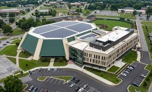 Aerial view of a large building with a green, multi-faceted roof covered in solar panels, adjacent to a modern office building.