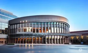 Modern circular building with glass facade and canopy entrance, reflecting lights on wet pavement under a clear sky.