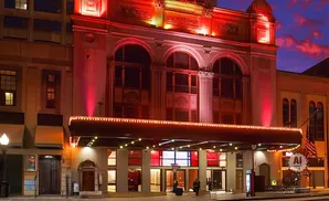 Exterior of a red-lit, ornate theater at dusk, with glowing marquee lights and a dramatic sky.