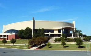 A large, modern, cream-colored arena with a curved facade, set against a clear blue sky, surrounded by green lawns and trees.
