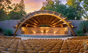 Outdoor amphitheater stage with rows of wooden seats under a curved wooden canopy, lit by warm lights.