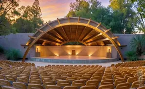 An outdoor amphitheater with a curved wooden roof, stage, and rows of empty wooden seats at sunset.