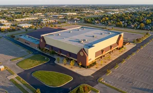 Aerial view of a large, light-yellow building with a dark roof, surrounded by empty parking lots and trees.