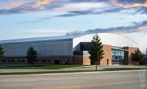 Exterior of a modern athletic facility with a large arched roof, brick accents, and glass entrance.