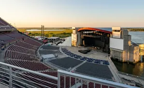 Outdoor amphitheater with a stage on a body of water, set against a marshy landscape and clear sky at sunset.