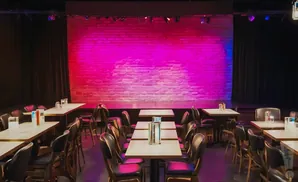 A comedy club stage with a brick wall backdrop bathed in pink and purple light. Tables and chairs are set up in front of the stage.