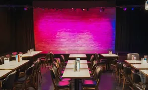 Empty comedy club with a brick stage lit pink and blue, and tables and chairs in the foreground.