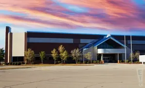A modern building with a large, angled blue roof and a red brick facade under a colorful sunset sky.