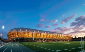 Modern stadium with a glowing, metallic, oval-shaped facade, lit by stadium lights at dusk.