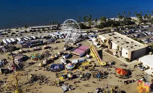 Aerial view of a busy seaside fair with rides, attractions, and a parking lot.