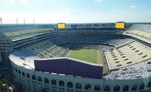Aerial view of a large, empty football stadium with yellow and blue seats, a green field, and two large scoreboards.
