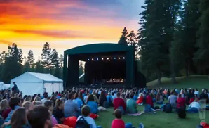 An outdoor concert with a large audience sitting on the grass as the sun sets behind a stage.