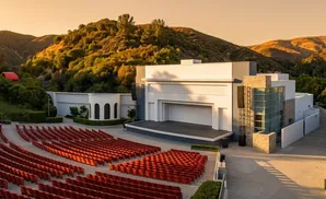 Outdoor amphitheater with red seats facing a stage, set against a backdrop of rolling hills at sunset.