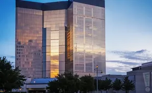 Tall glass skyscraper reflects sunset, with trees and a lamp post in the foreground.